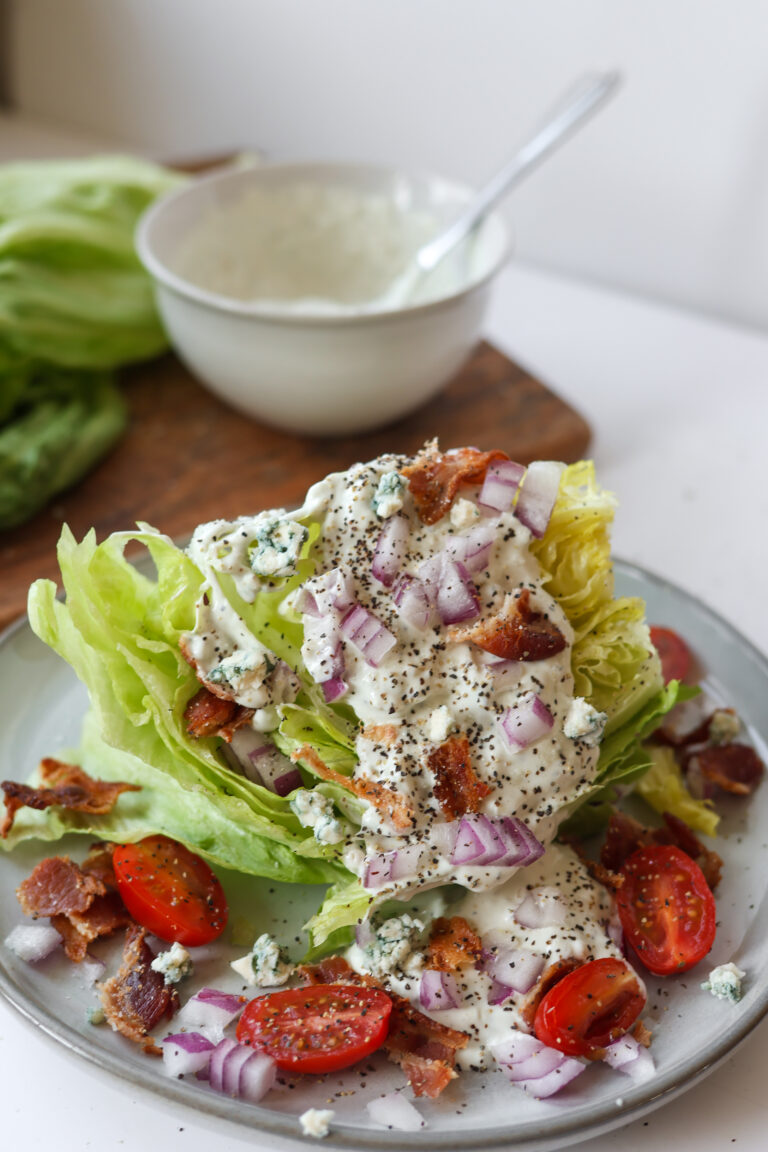 Blue cheese wedge salad topped with crispy bacon, cherry tomatoes, red onion, and creamy dressing on a white plate.