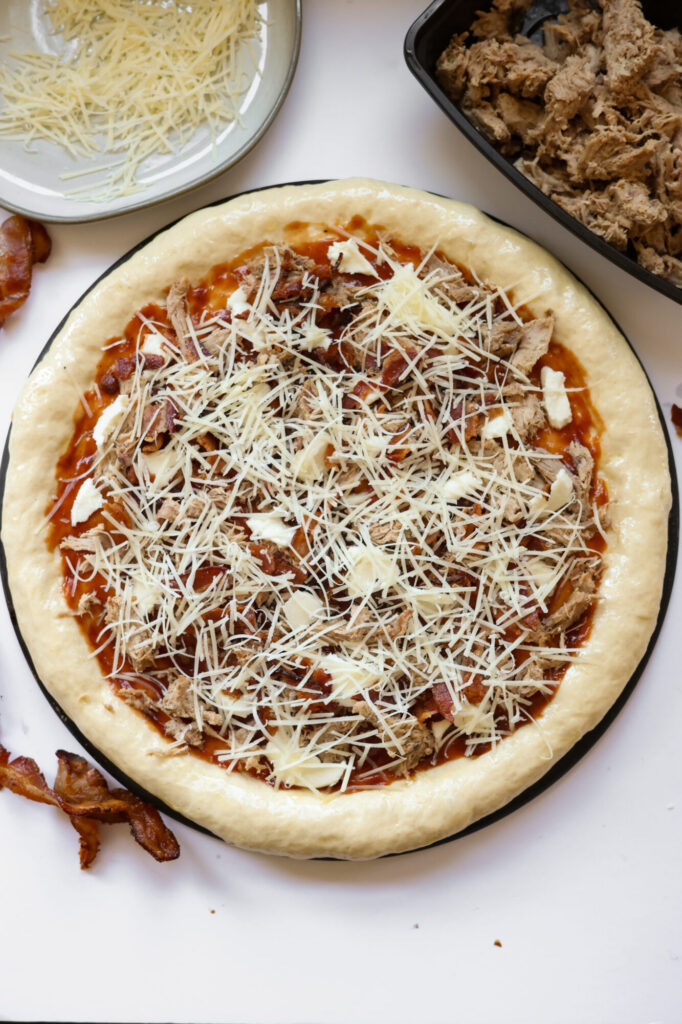 Homemade pizza dough topped with BBQ sauce, pulled pork, and cheeses before baking.