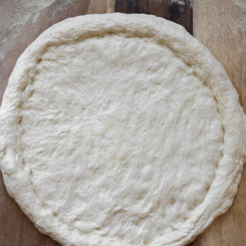 Shaped homemade pizza crust made from all-purpose flour dough on wooden board with olive oil and semolina flour, ready for toppings in an easy pizza dough recipe.