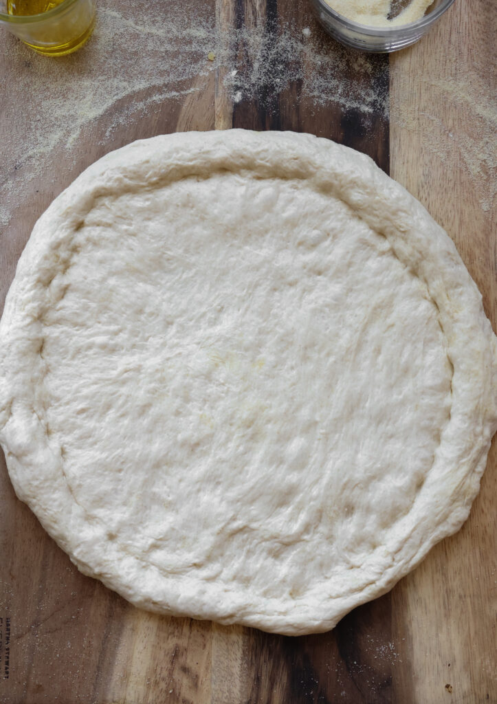Shaped homemade pizza crust made from all-purpose flour dough on wooden board with olive oil and semolina flour, ready for toppings in an easy pizza dough recipe.