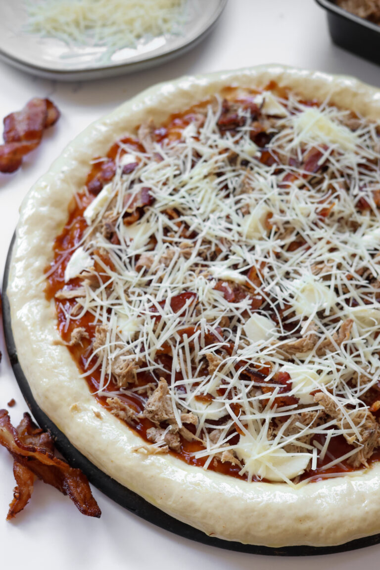 Unbaked pizza made with homemade all-purpose flour crust, layered with BBQ sauce, shredded mozzarella cheese, and fresh toppings, ready for baking into a chewy, crispy crust.