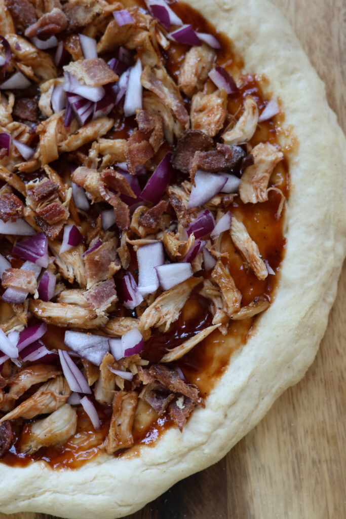 Uncooked BBQ chicken bacon pizza topped with shredded chicken breast, fresh mozzarella slices, crispy bacon bits, thinly sliced red onions, and parmesan cheese on homemade pizza dough ready for baking