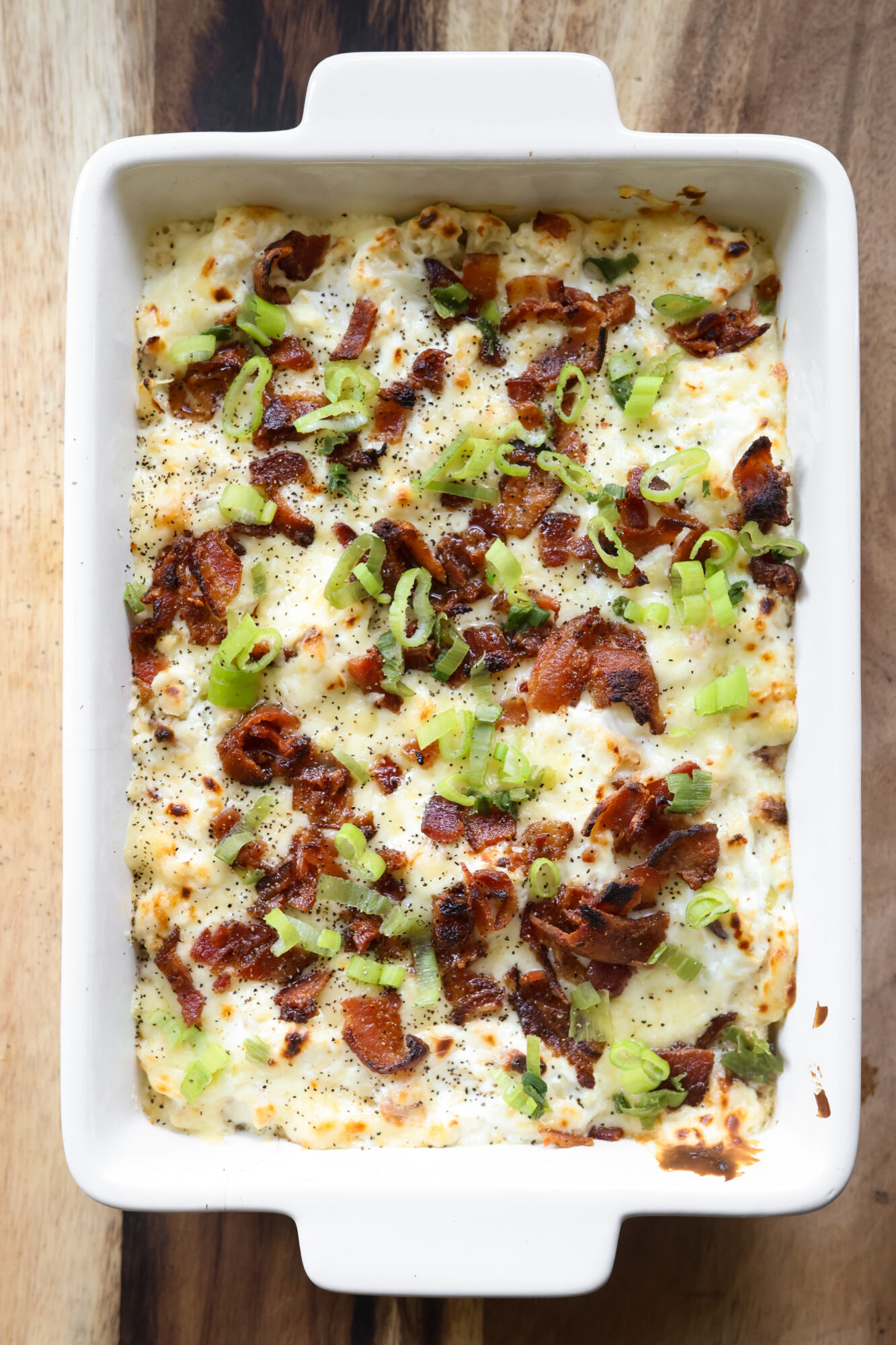 Creamy loaded cauliflower casserole with melted sharp cheddar cheese and crispy bacon on top in a white casserole dish.