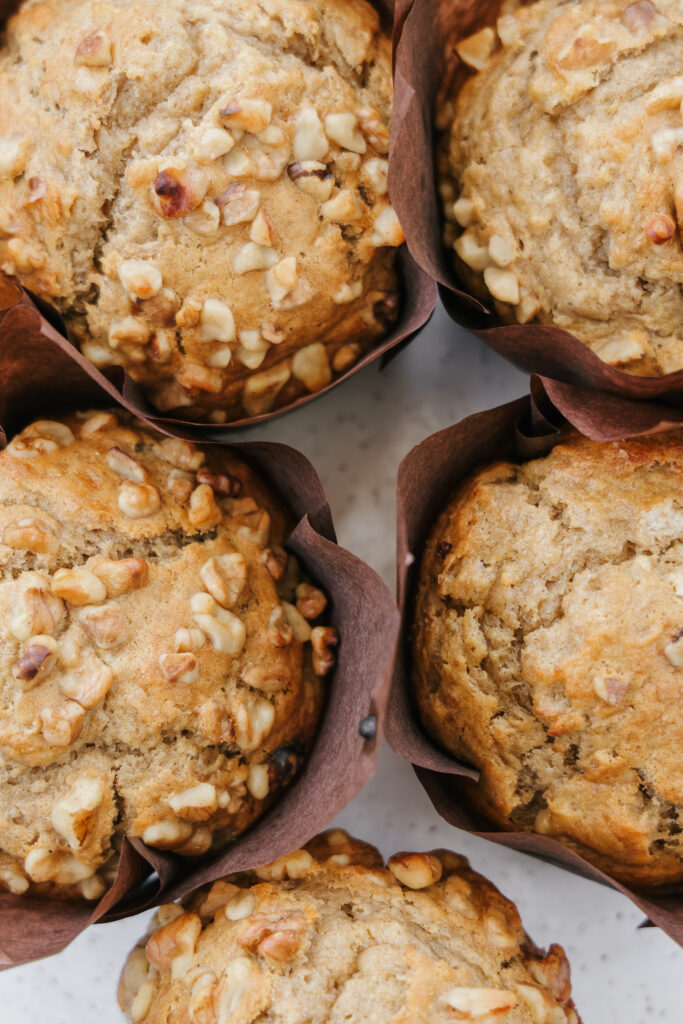 Jumbo banana muffins with sour cream in brown parchment tulip wrappers topped with chopped walnuts on a white marble surface