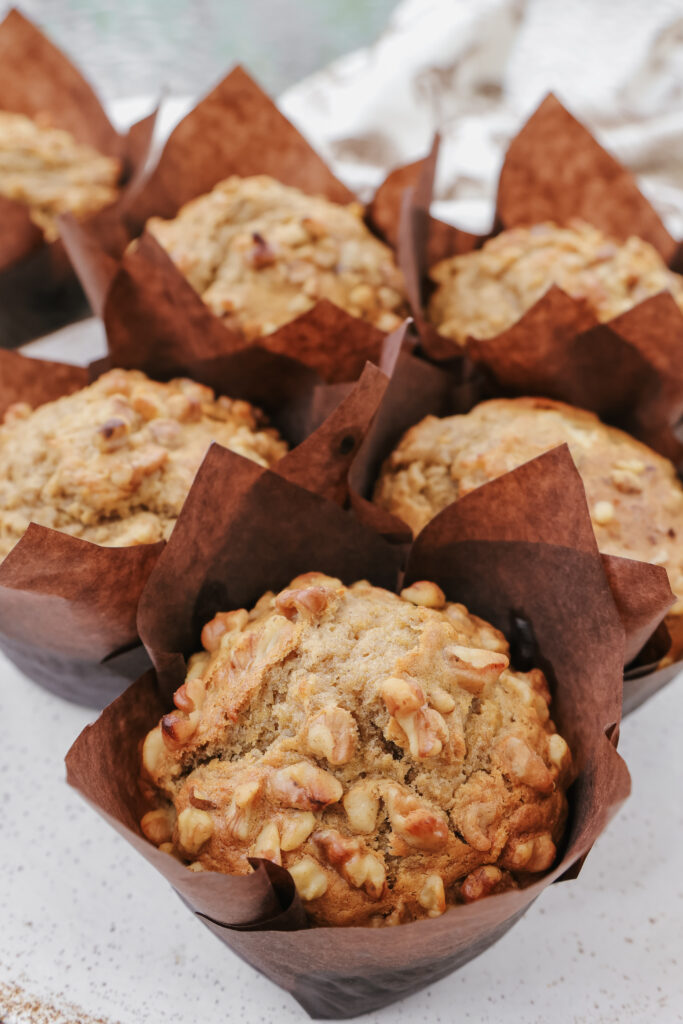 Six bakery style banana muffins with sour cream and chopped walnuts in brown tulip parchment liners on white marble