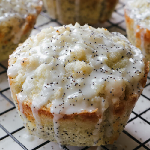 Single jumbo lemon poppy seed muffin with streusel topping and lemon glaze on a wire rack, with more muffins in tulip liners visible in the background.