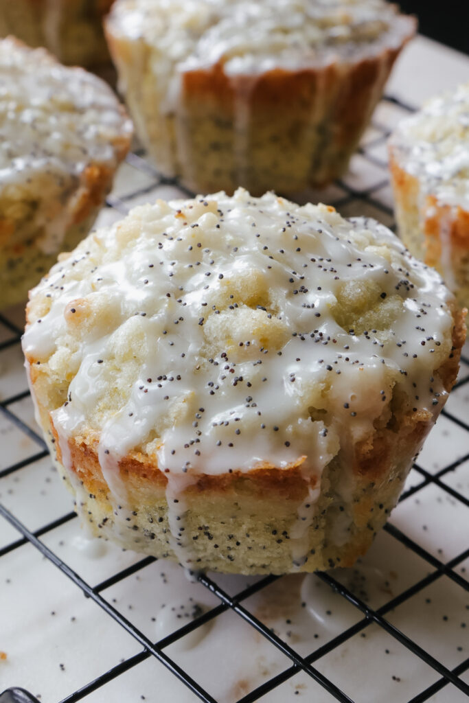 Single jumbo lemon poppy seed muffin with streusel topping and lemon glaze on a wire rack, with more muffins in tulip liners visible in the background.