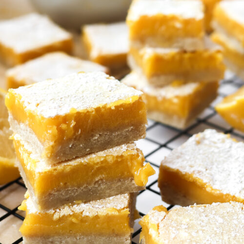 Lemon bars with shortbread crust stacked on a wire rack with fresh lemons in the background.