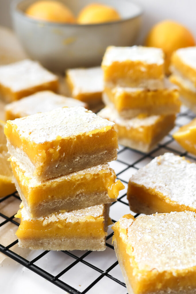 Lemon bars with shortbread crust stacked on a wire rack with fresh lemons in the background.