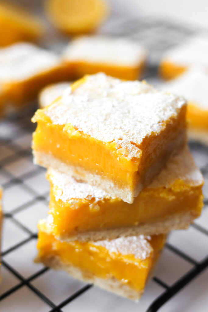 Lemon bars with shortbread crust stacked on a wire cooling rack dusted with powdered sugar.