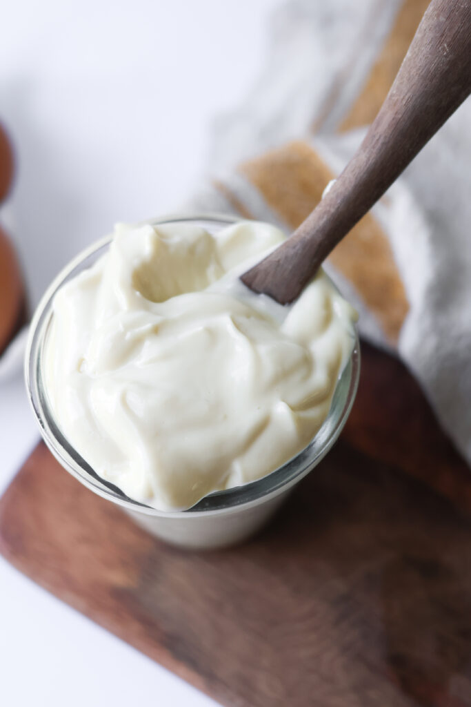 Overhead view of homemade mayonnaise with avocado oil in a glass jar on a wooden cutting board with a spoon and brown eggs nearby.