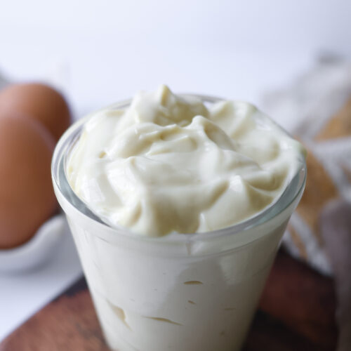 A glass jar filled with thick, creamy homemade avocado oil mayonnaise on a wooden board with brown eggs in the background.