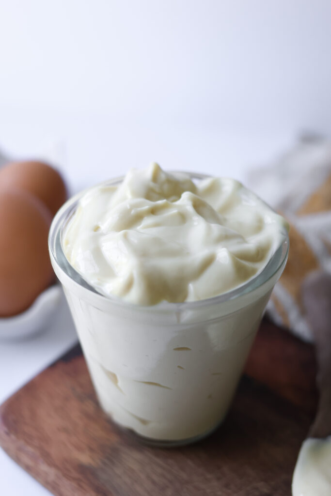 A glass jar filled with thick, creamy homemade avocado oil mayonnaise on a wooden board with brown eggs in the background.
