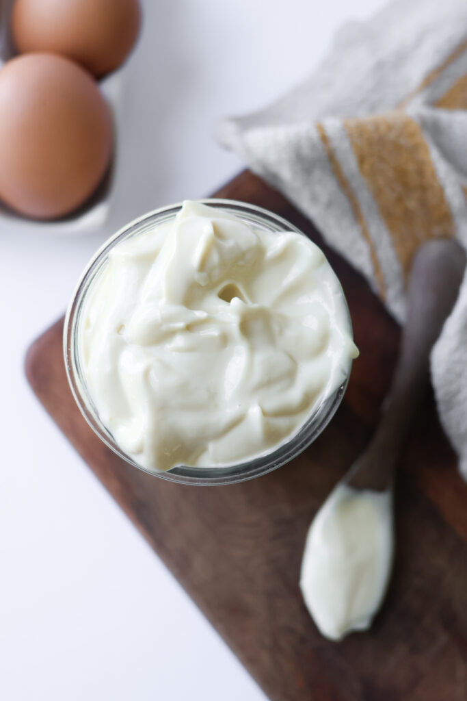 Close-up of homemade avocado oil mayo in a glass jar with a wooden spoon resting inside on a wooden cutting board.