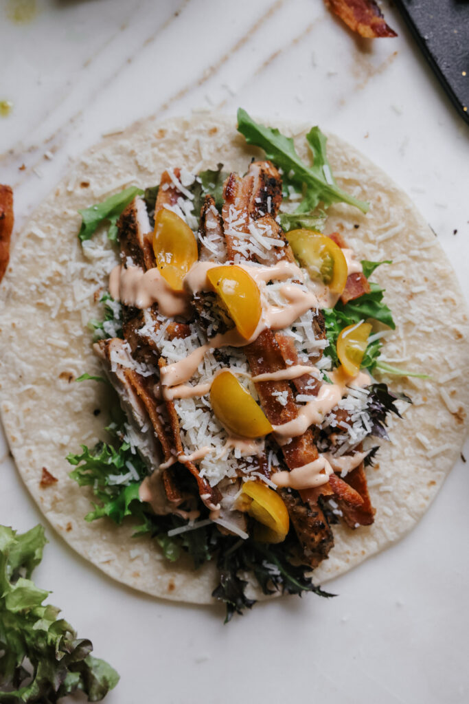 Open-faced tortilla wrap with grilled chicken, bacon, greens, cherry tomatoes, parmesan, and sriracha mayonnaise drizzled on top