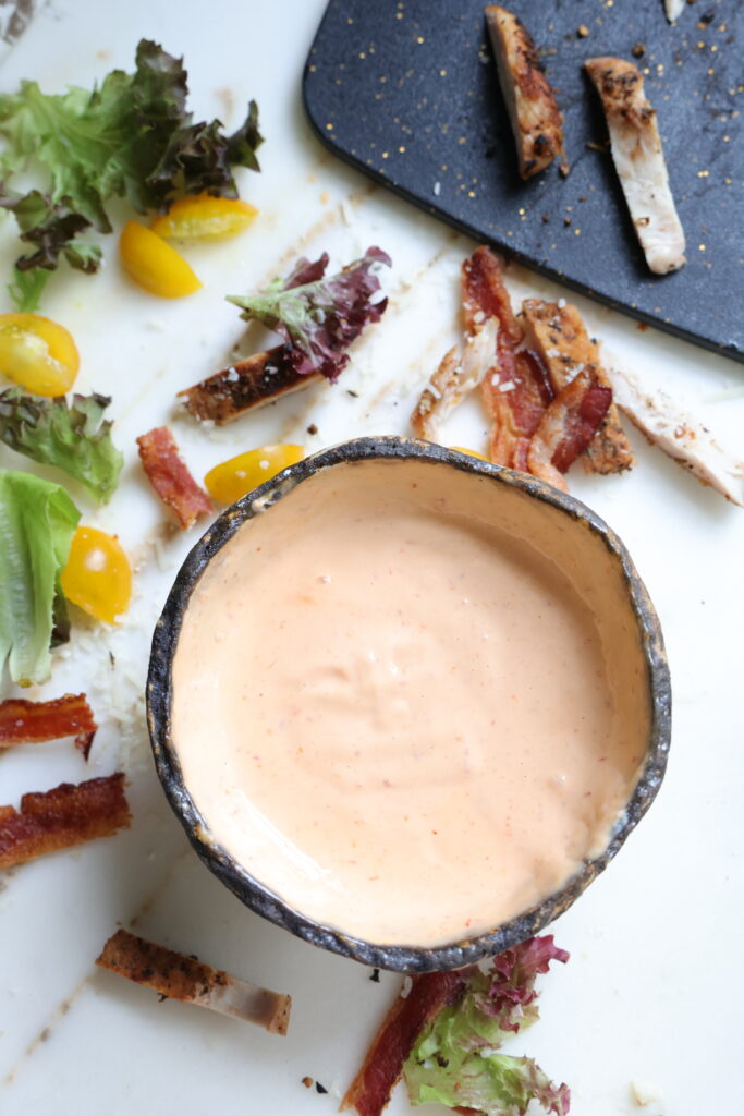 Bowl of creamy sriracha mayonnaise surrounded by bacon, grilled chicken, lettuce, and cherry tomatoes.
