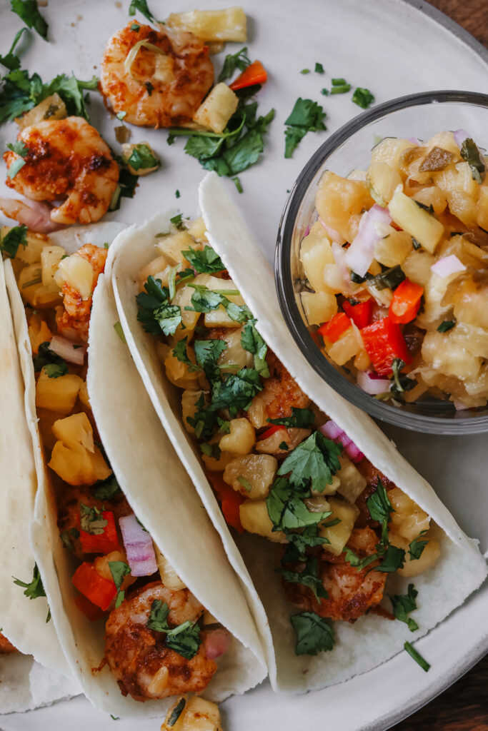 Overhead shot of shrimp tacos with pineapple salsa on a plate next to a bowl of fresh pineapple salsa