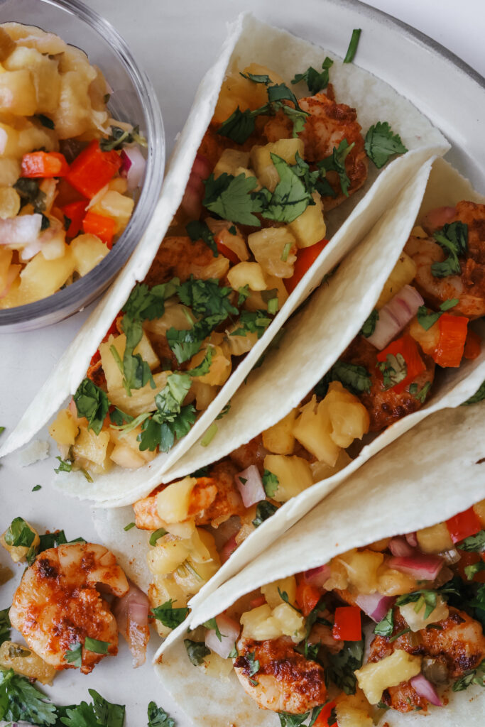 Shrimp tacos with pineapple salsa filling spilling out of tortillas next to a bowl of salsa