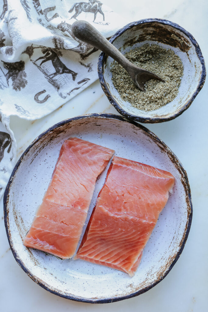 Two raw salmon fillets on a plate next to a bowl of brown sugar spice rub with a wooden spoon