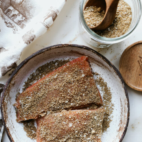 Raw salmon fillets coated in brown sugar salmon rub on a speckled plate beside a jar of rub