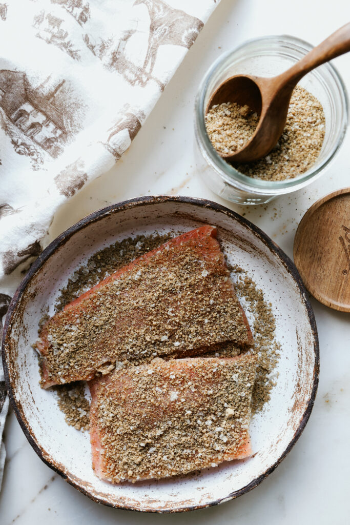 Raw salmon fillets coated in brown sugar salmon rub on a speckled plate beside a jar of rub